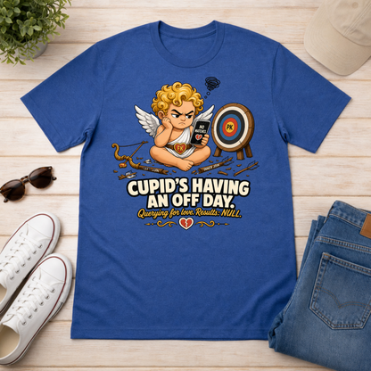 A royal blue graphic t-shirt laid flat on a light wood surface, surrounded by lifestyle props including white sneakers, folded blue jeans, sunglasses, a plant, and a beige cap. The t-shirt features a cartoon cupid sitting at a desk with a phone, bow and arrow, and a target, with the text 'CUPID'S HAVING AN OFF DAY Quirking for her Romance NIKEL'.